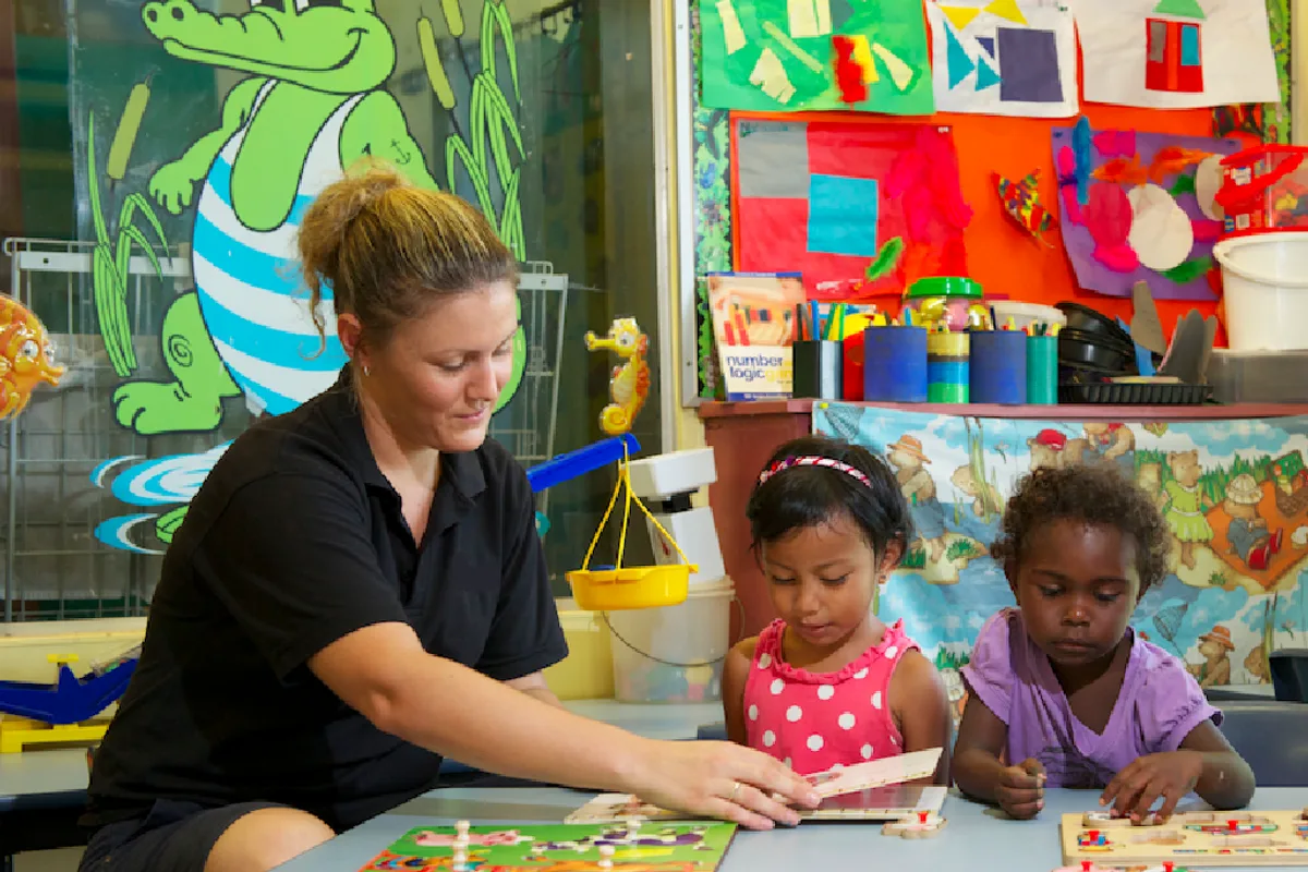 A support worker sharing puzzle boards with two children in a colourful classroom